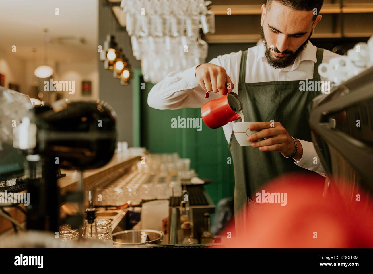 In einem warmen und einladenden Café gießt ein engagierter Barista fachmännisch Milch in eine Kaffeetasse und sorgt so für ein angenehmes Erlebnis für Gäste. Stockfoto