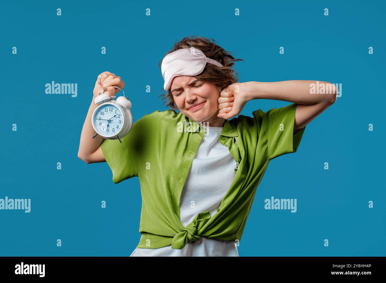 Geweckt von Wecker, eine verschlafene junge Frau, die es in der Hand hält. Blauer Hintergrund. Früh um 6 Uhr morgens. Die faule Dame hat nicht genug geschlafen, Konzept Stockfoto