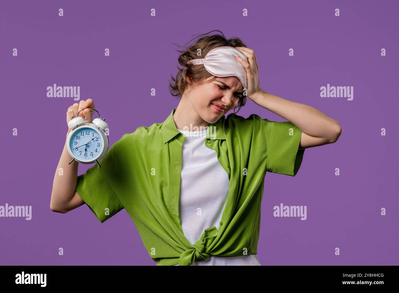 Geweckt von Wecker, eine verschlafene junge Frau, die es in der Hand hält. Violetter Hintergrund. Früh um 6 Uhr morgens. Die faule Dame hat nicht genug geschlafen Stockfoto