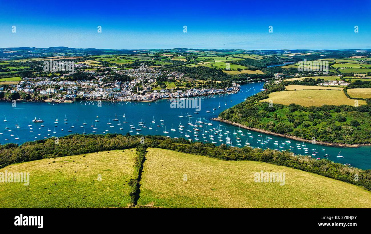 Blick aus der Vogelperspektive auf eine malerische Küstenstadt, umgeben von üppigen grünen Hügeln und einem Fluss voller Segelboote. Der klare blaue Himmel unterstreicht die lebendige Atmosphäre Stockfoto