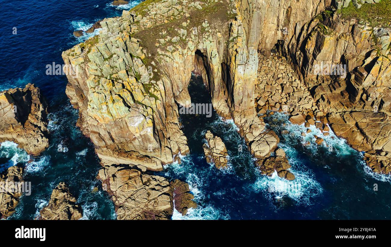 Blick aus der Vogelperspektive auf zerklüftete Küstenklippen mit einem natürlichen Felsbogen, umgeben von tiefblauem Meer. Wellen krachen gegen die felsige Küste und schaffen Stockfoto