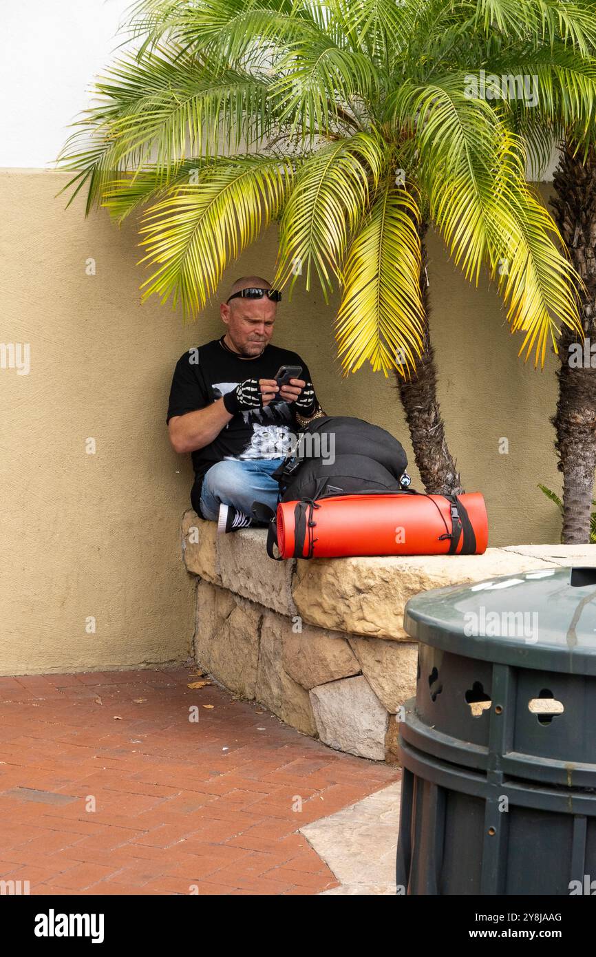 Ein Vielreisender hält an und ruht sich mit seiner Bettrolle und seinem Rucksack auf einer Steinmauer an der State Street in Santa Barbara, Kalifornien aus. Stockfoto