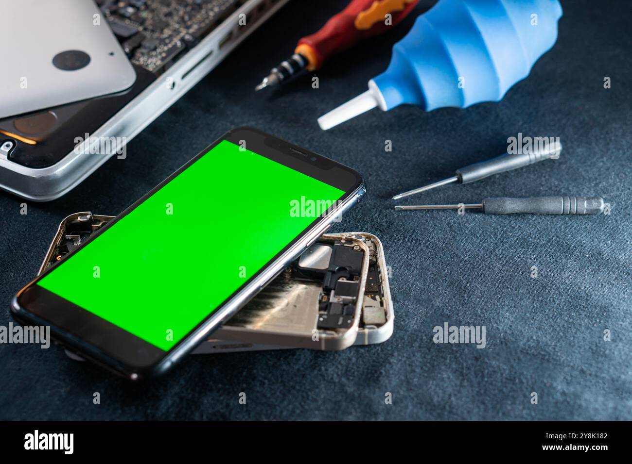 Ein Smartphone mit grünem Bildschirm wird auf einem zerlegten Telefon platziert. Zu den umgebenden Werkzeugen gehören ein Schraubendrehersatz, ein Gebläse und ein Laptop im Stockfoto