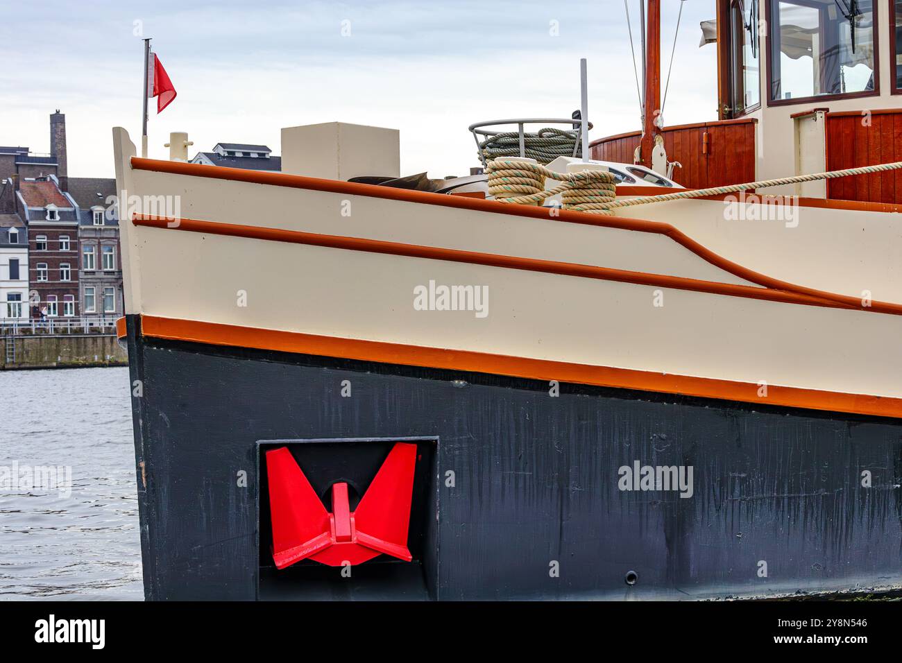 Nahaufnahme des Bugs eines Schiffes, das auf der Maas fährt, roter Anker, rote Flagge, Seile, Flybridge, Gebäude im Hintergrund, bewölkter Tag in Maastricht, South Lim Stockfoto