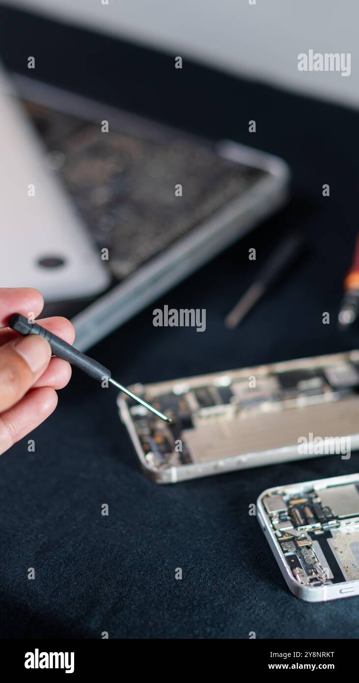 Nahaufnahme einer Person, die ein Smartphone mit einem Schraubendreher repariert, mit einem Laptop im Hintergrund. Stockfoto