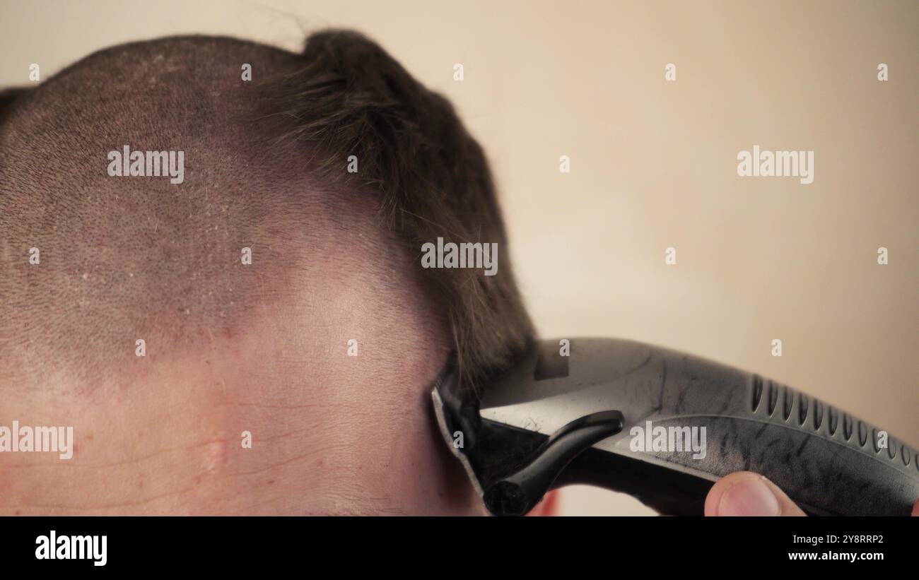 Gutaussehender Mann im Badezimmer rasiert sich seinen Kopf mit einem elektrischen Rasierer aufgrund der Coronavirus-Quarantäne-Selbstisolierung. Teil des Kopfes im Rahmen. Schließen Stockfoto