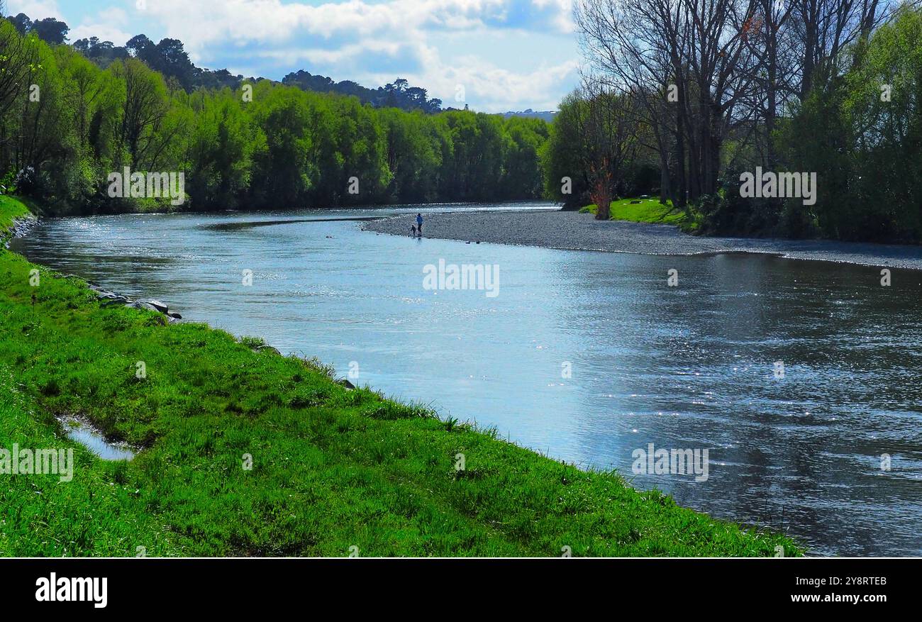 Langsam fließender Hutt River, Neuseeland, mit einem Kind und einem Erwachsenen, die auf einem niedrigen Steinufer an einer Flussbiegung stehen. Üppiges Frühlingswachstum an beiden Ufern Stockfoto