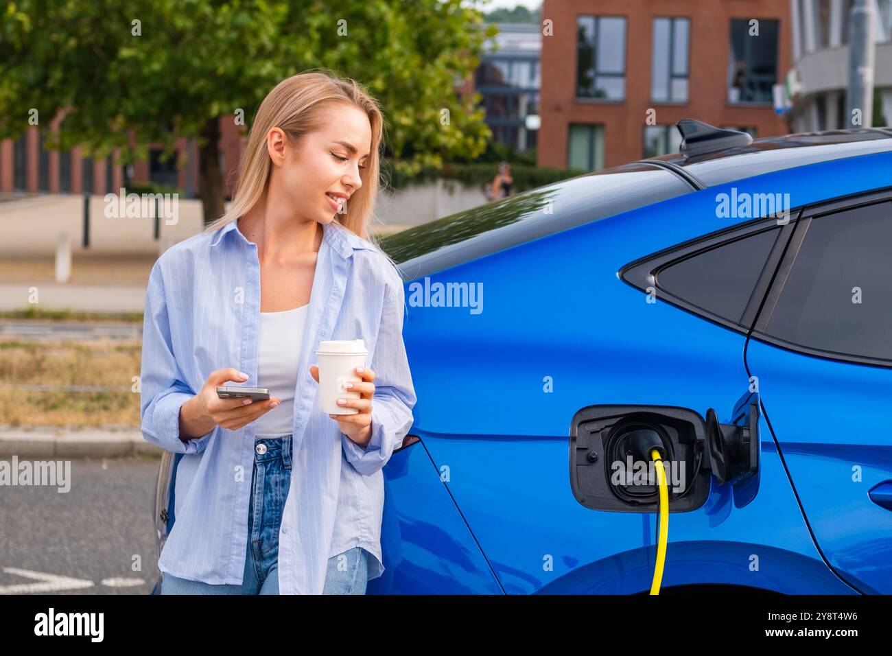 Die blonde Frau genießt Kaffee, während sie ihr Elektroauto auflädt und den Akkustand mit dem Smartphone an einer modernen Station überprüft. Stockfoto