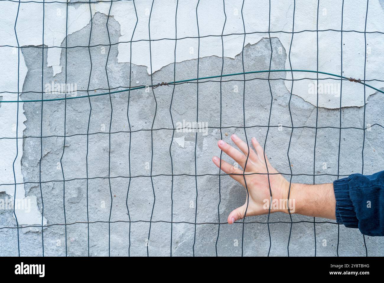 Die Hand hinter dem Zaun gegen die verwitterte Mauer. Rechtzeitig erfasst. Status Quo. Stockfoto