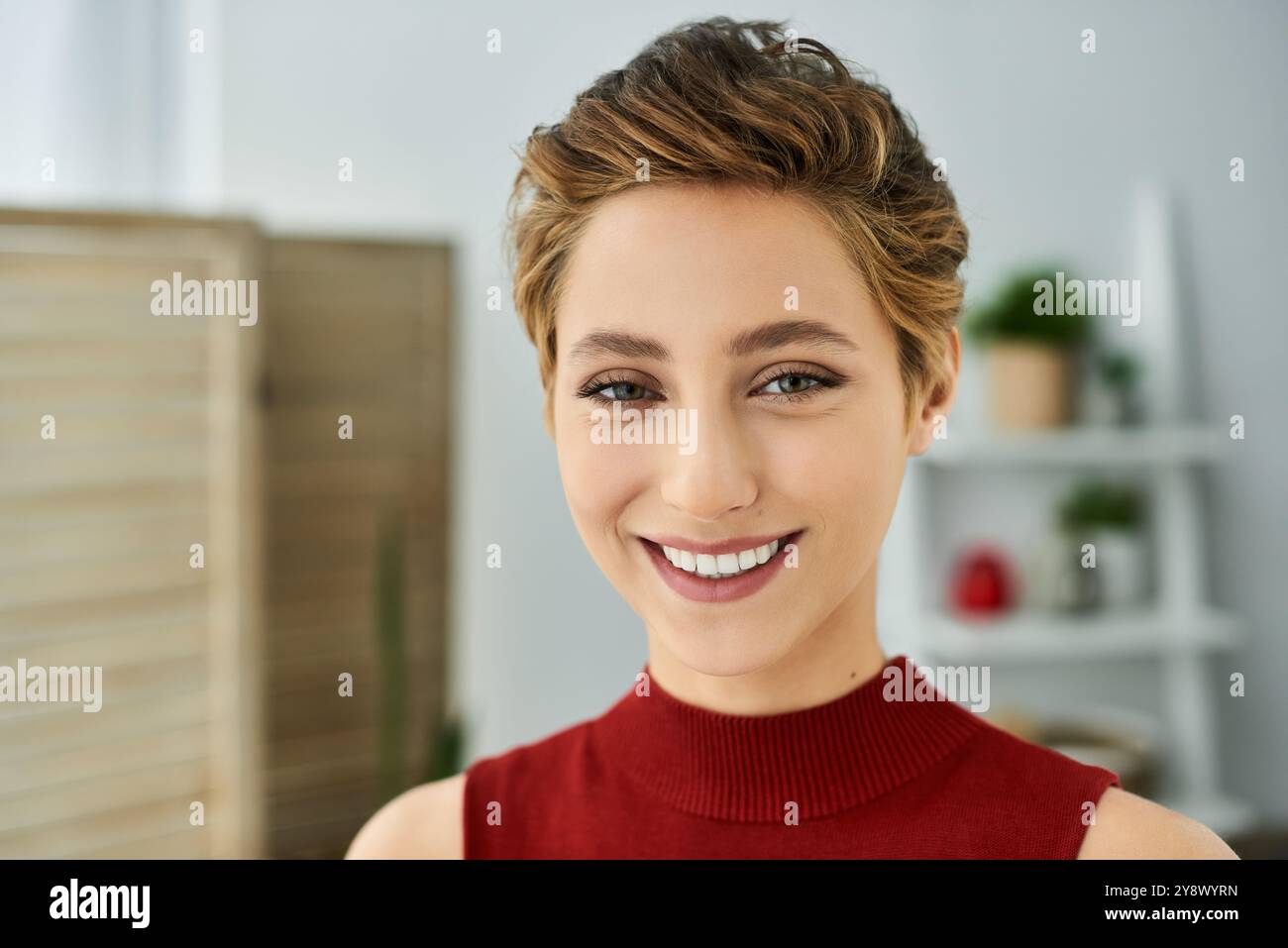 Eine junge Frau mit kurzen Haaren lächelt strahlend in ihrem roten Tanktop und genießt einen entspannten Tag zu Hause. Stockfoto