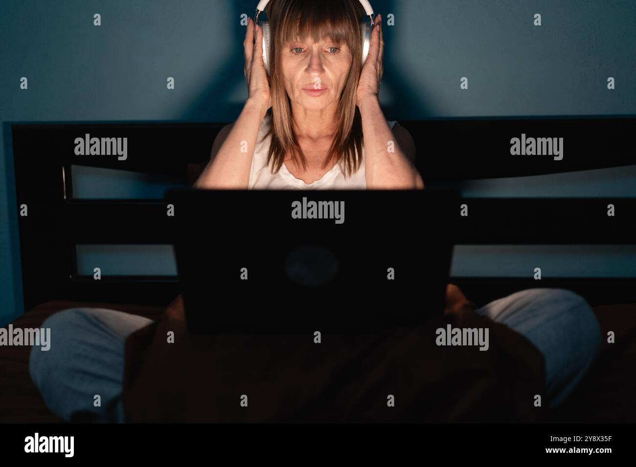 Eine konzentrierte Frau, die Kopfhörer trägt, benutzt einen Laptop, während sie nachts auf dem Bett sitzt und Überstunden macht. Der schwach beleuchtete Raum unterstreicht ihre Hingabe und ihren Konzent Stockfoto