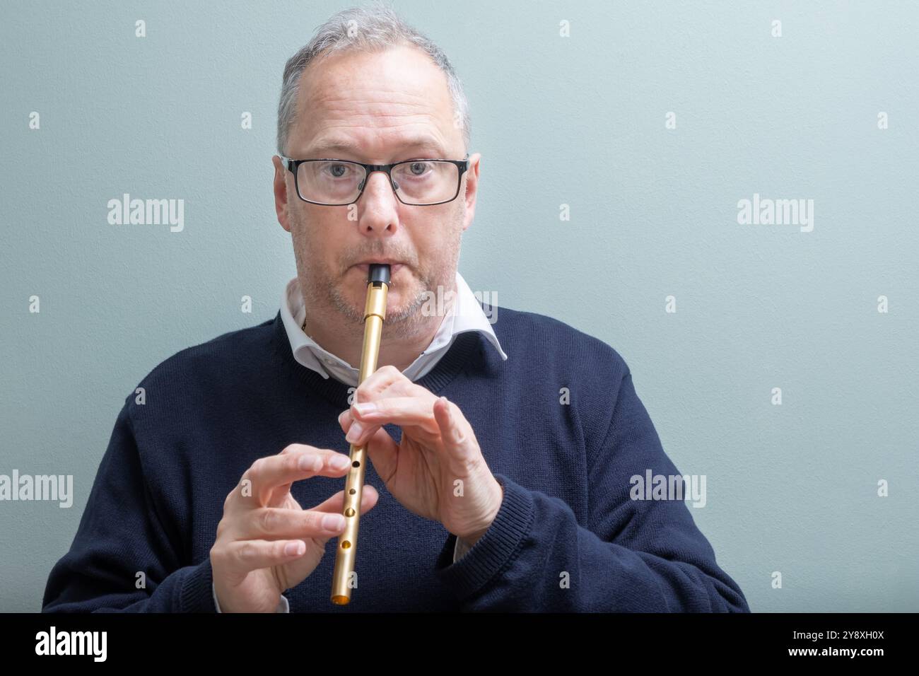 Mann mit grauen Haaren, lässig gekleidet, mit dunkler Brille, spielt eine goldfarbene Blechpfeife, Pennypfeife, traditionelle irische Volksmusik. Stockfoto