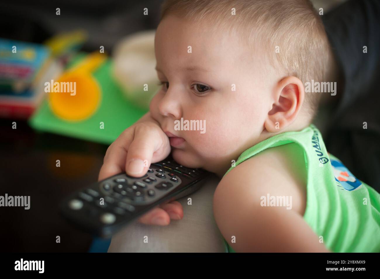 Das kleine Kind liegt auf einer Couch und legt sein Kinn auf eine Hand, während es eine Fernbedienung packt. Der lebhafte Spielbereich im Hintergrund sorgt für ein Playfu Stockfoto