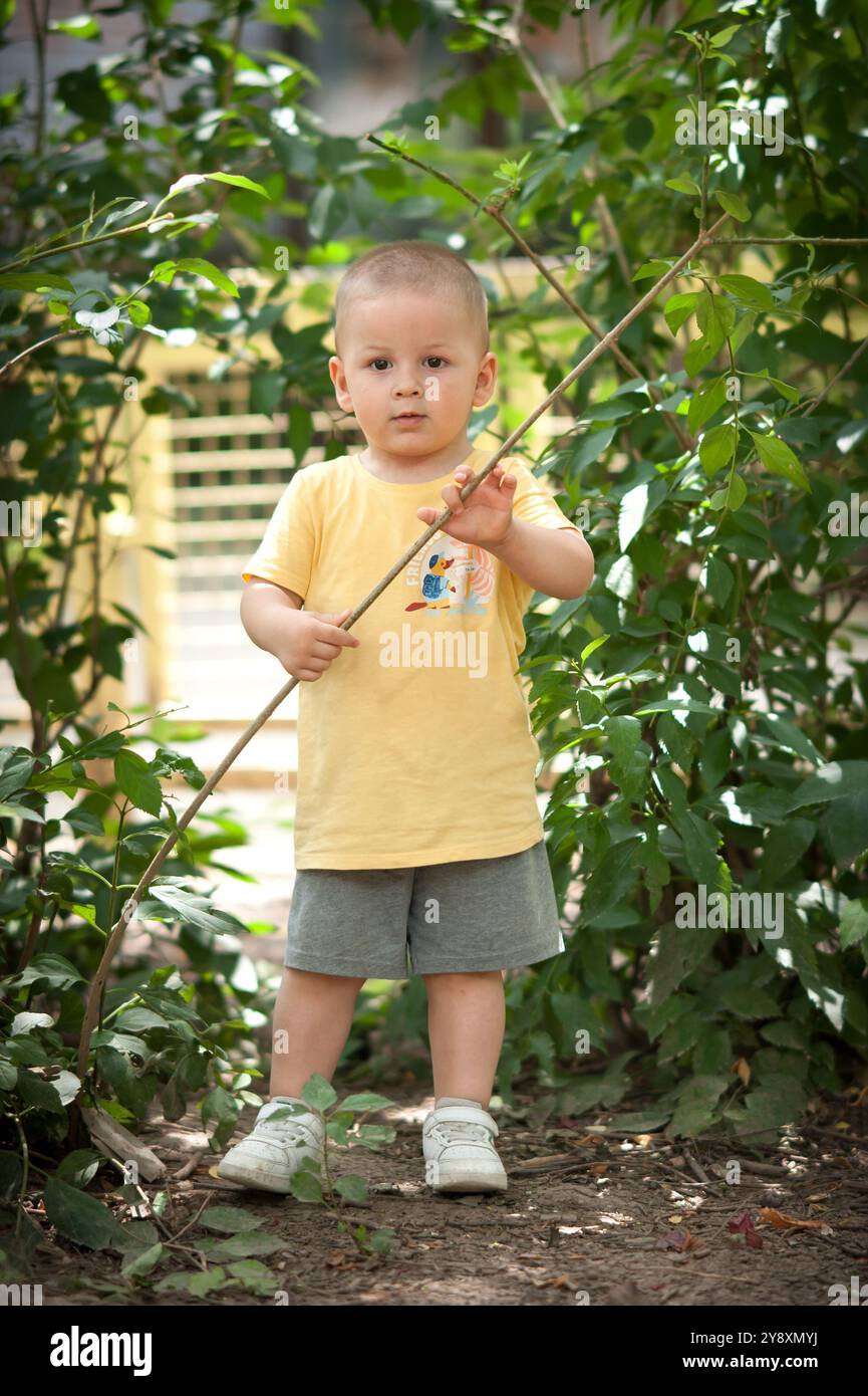Das Kleinkind steht in einem lebendigen Garten und hält einen Stock, während es von dichten grünen Blättern umgeben ist. Das Kind trägt ein gelbes Hemd und Shorts, die Unschuld verkörpern Stockfoto