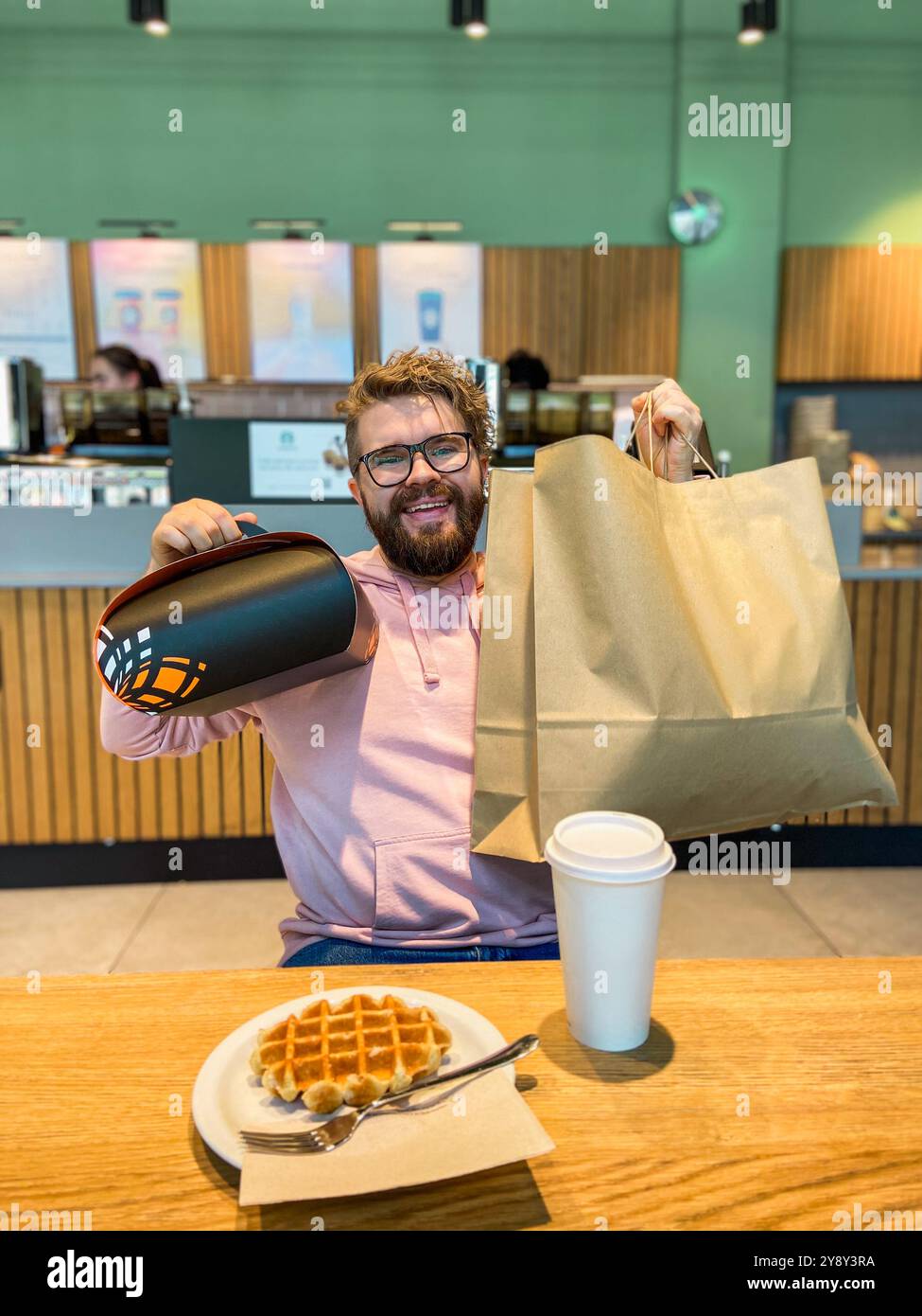 Ein überraschter Mann sitzt mit einer Waffel an einem Café-Tisch, hält eine große Einkaufstasche und eine Geschenkbox zum Mitnehmen. Moment der unerwarteten Freude und des Genusses in einem Stockfoto