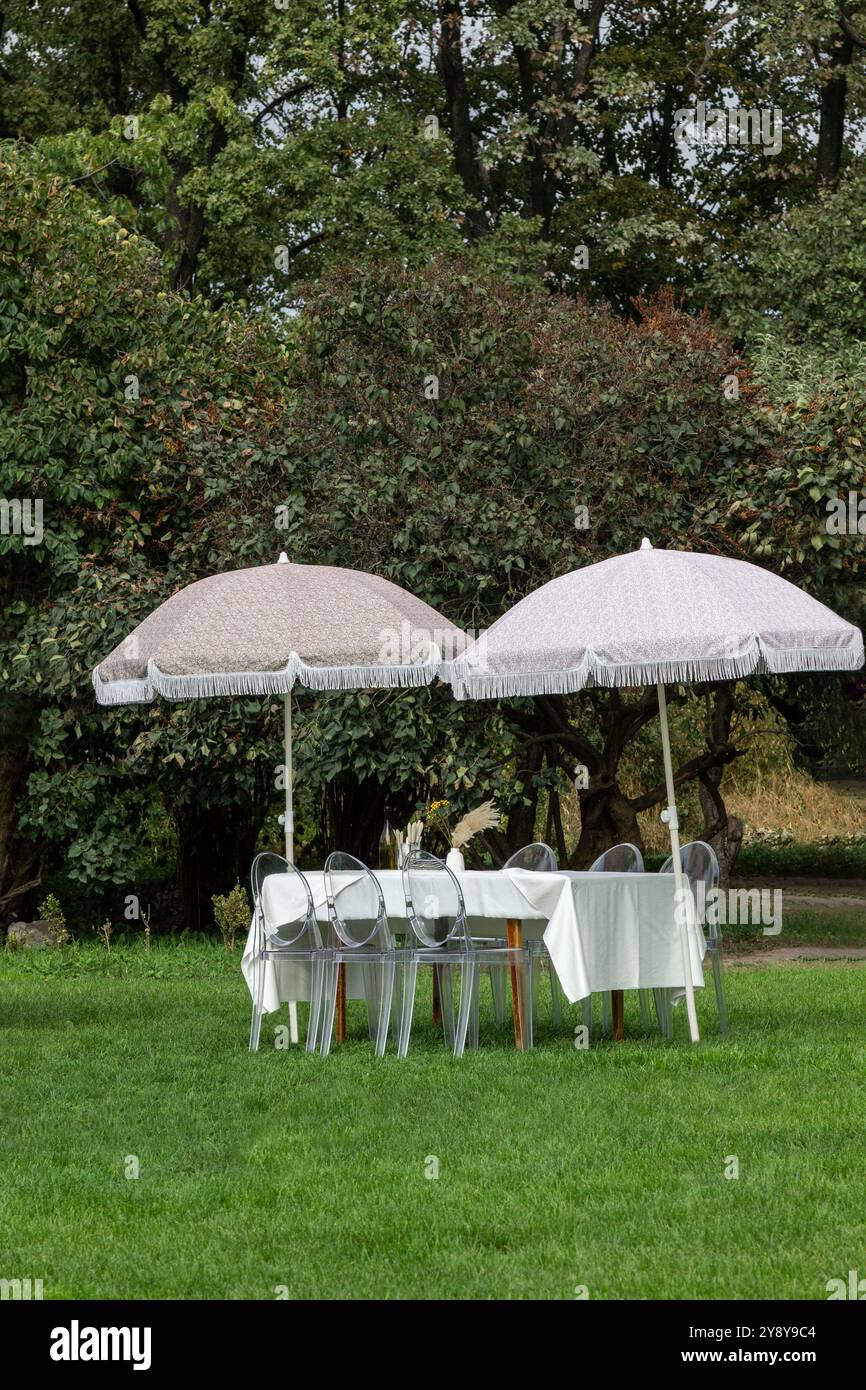 Ein Tisch auf einem grünen Rasen. Zwei Sonnenschirme in der Nähe des Tisches. Der Tisch ist mit einer weißen Tischdecke bedeckt. Stockfoto