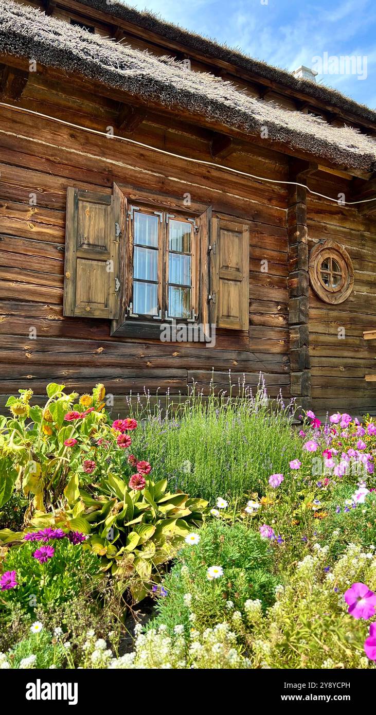 Vertikales Foto eines Fensters aus einem alten Holzhaus mit einem strohbedeckten Dach. Großes Blumenbeet im Vordergrund. Rustikale Architektur, Landschaft, t - Smartphone-aufgenommenes Stockfoto