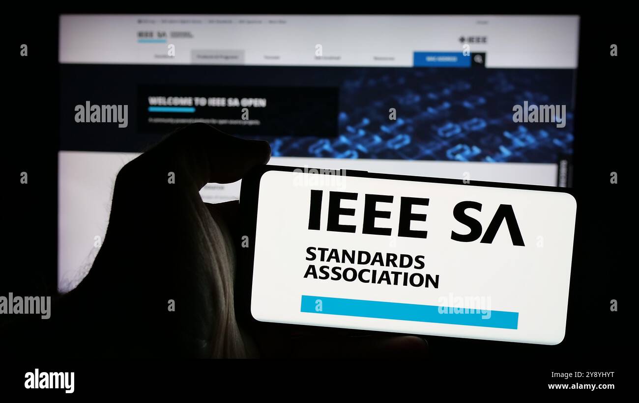 Person, die ein Mobiltelefon mit dem Logo der IEEE Standards Association auf dem Bildschirm vor der Webseite hält. Schwerpunkt auf dem Display des Telefons. Stockfoto