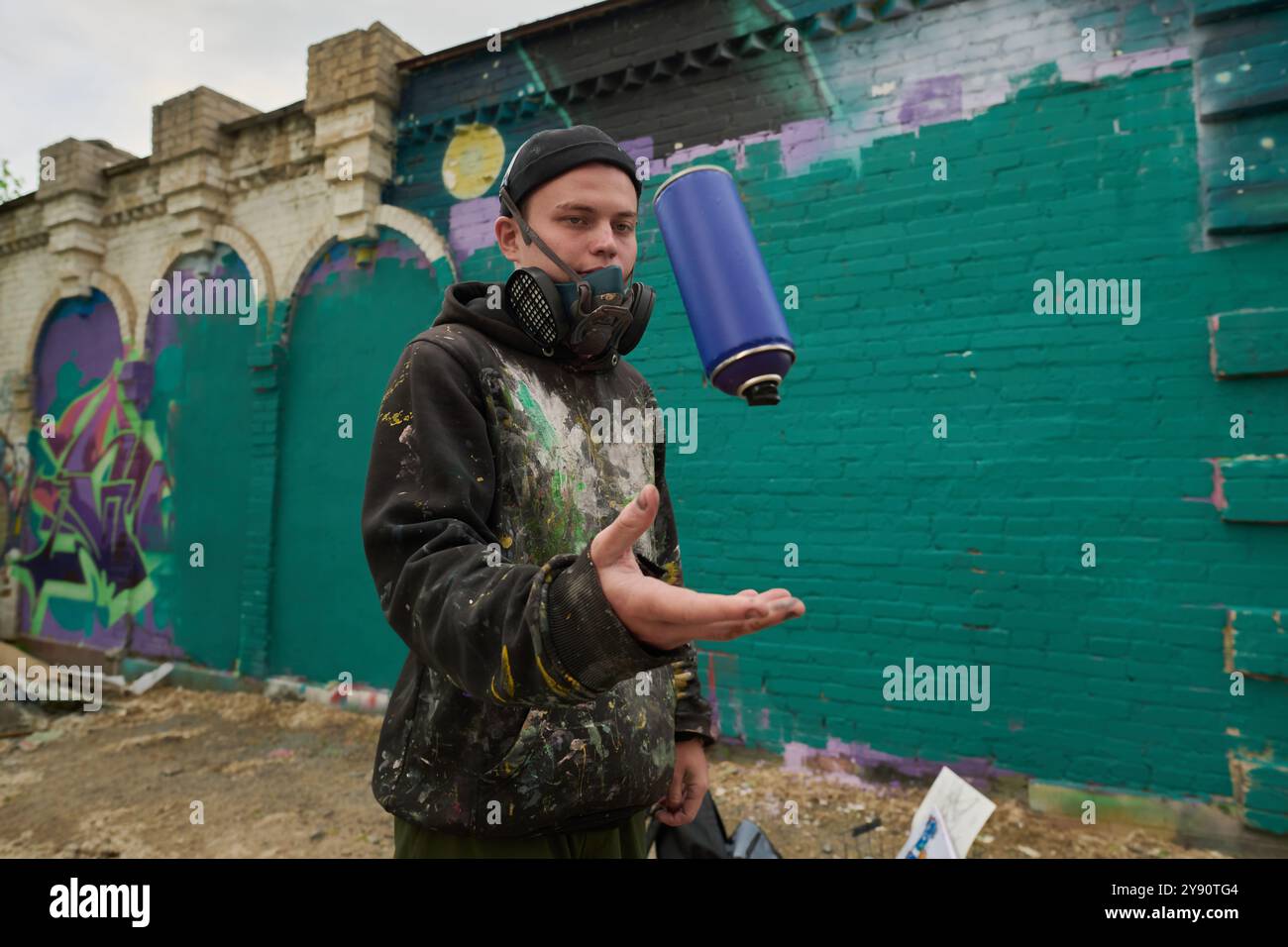 Ein Typ in Atemschutzmaske, Mütze und Hoodie wirft blaue Dose mit Farbe, während er gegen die grüne Wand des Gebäudes steht Stockfoto