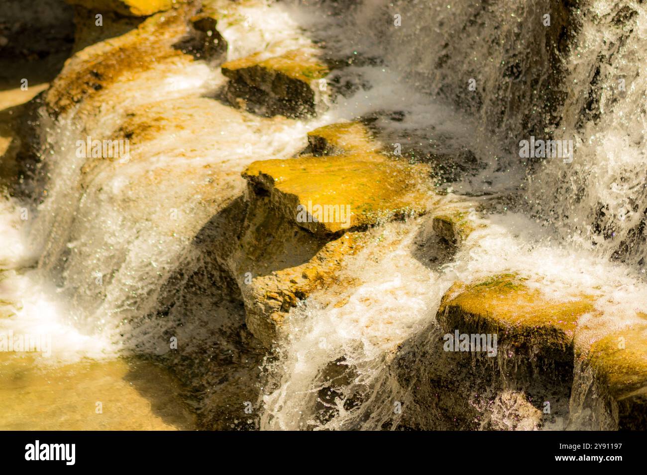 Ein kleiner Wasserfall, der über zerklüftete Felsen kaskadiert. Das Bild gleicht die warmen Erdtöne der Felsoberfläche mit dem kühlen, schaumigen Rauschen des Wassers aus. Stockfoto
