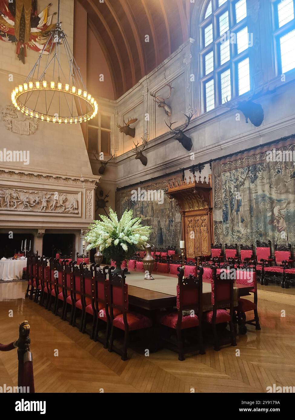 Ein großer Speisesaal mit einem langen Holztisch, umgeben von kunstvollen Stühlen. Der Raum ist mit großen Wandteppichen und aufgehängten Tierköpfen geschmückt Stockfoto