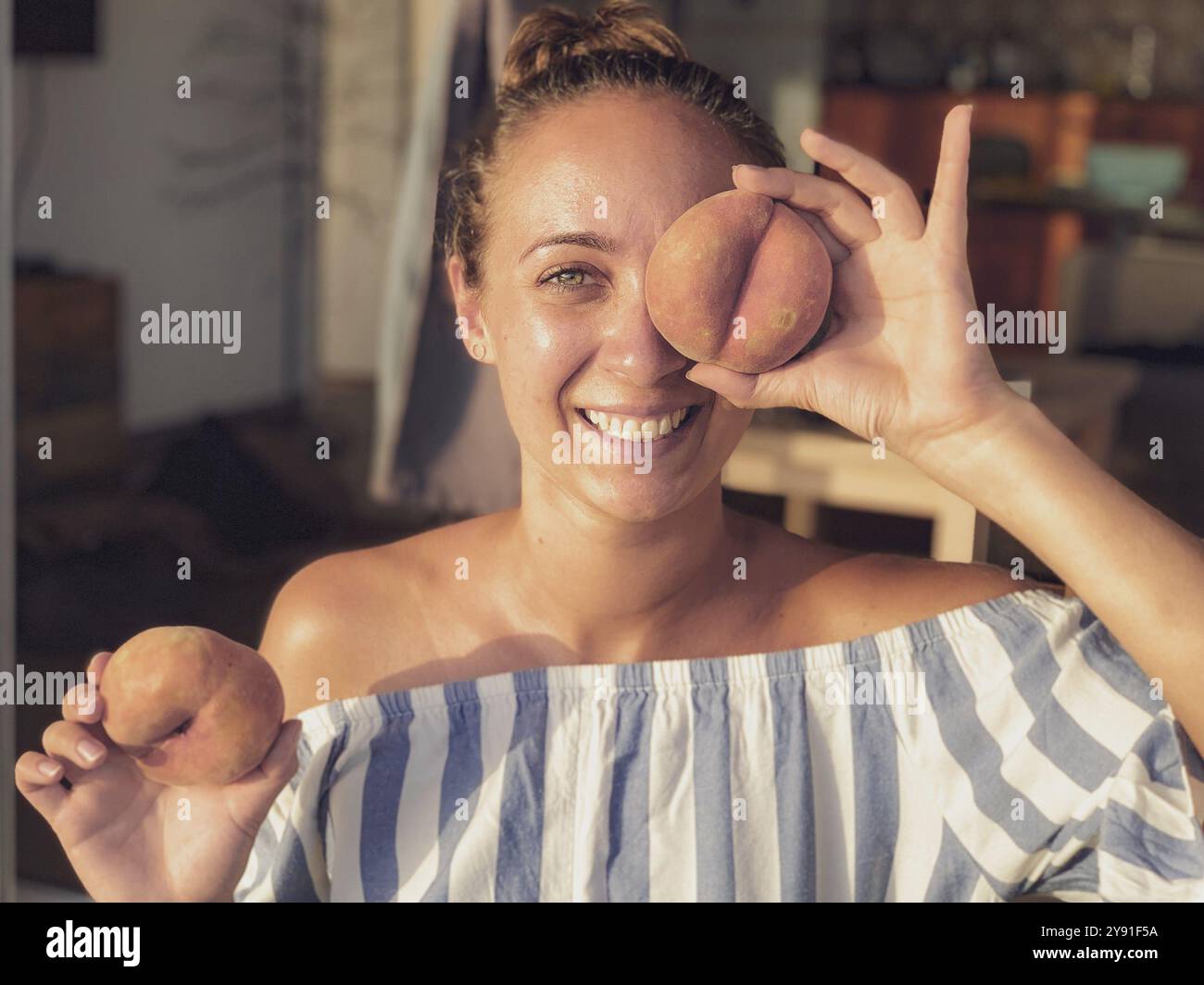 Lachende Frau, die einen Pfirsich vor einem Auge in einer gemütlichen Einrichtung hält Stockfoto