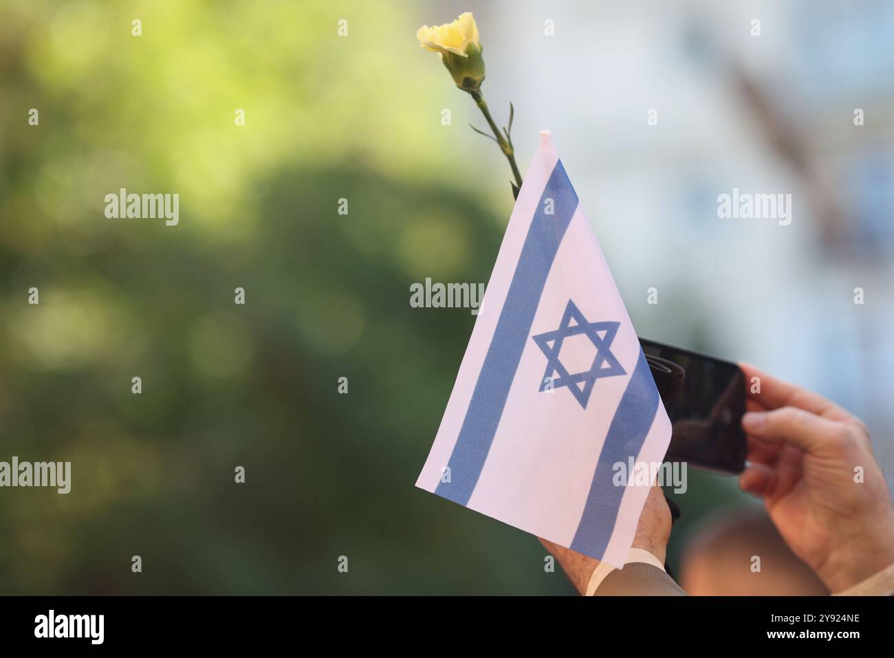 Details mit den Händen eines Mannes, der die israelische Flagge und eine Blume während einer Zeremonie zum Gedenken an die Angriffe vom Oktober 2023 hält. Stockfoto