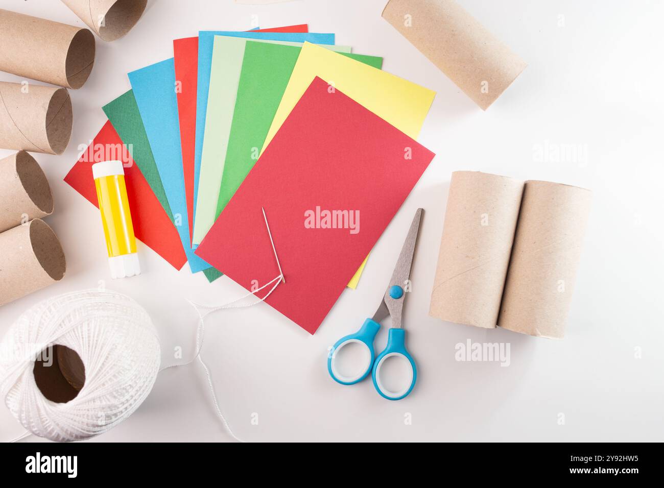 Bastelmaterialien für Papierdekorationen, mit farbigen Papierbögen, Pappröhrchen, Klebestift, Schere, Nadel, und weißes Gewinde. Flache Lay-Komposition für Heimwerker und Bastelprojekte. Stockfoto