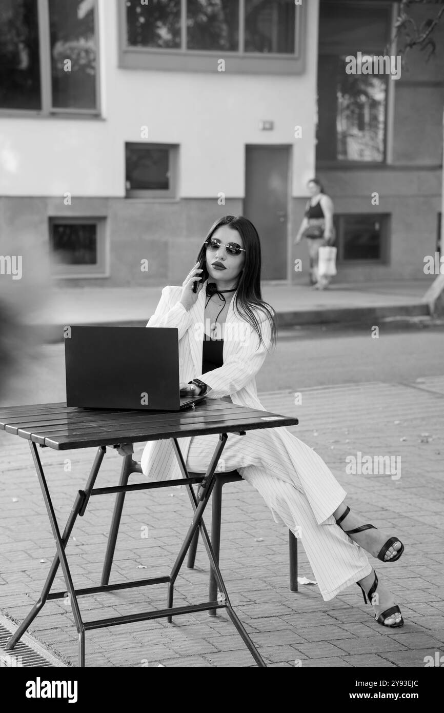 Eine stilvolle Geschäftsfrau, die im Freien an einem Laptop arbeitet, telefoniert. Stockfoto