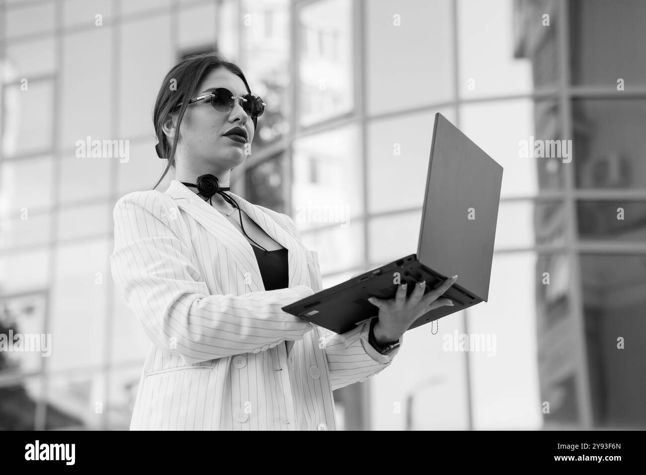 Stilvolle Geschäftsfrau mit Sonnenbrille in einem modernen urbanen Ambiente, die in einem Nadelstreifenanzug einen Laptop hält. Stockfoto