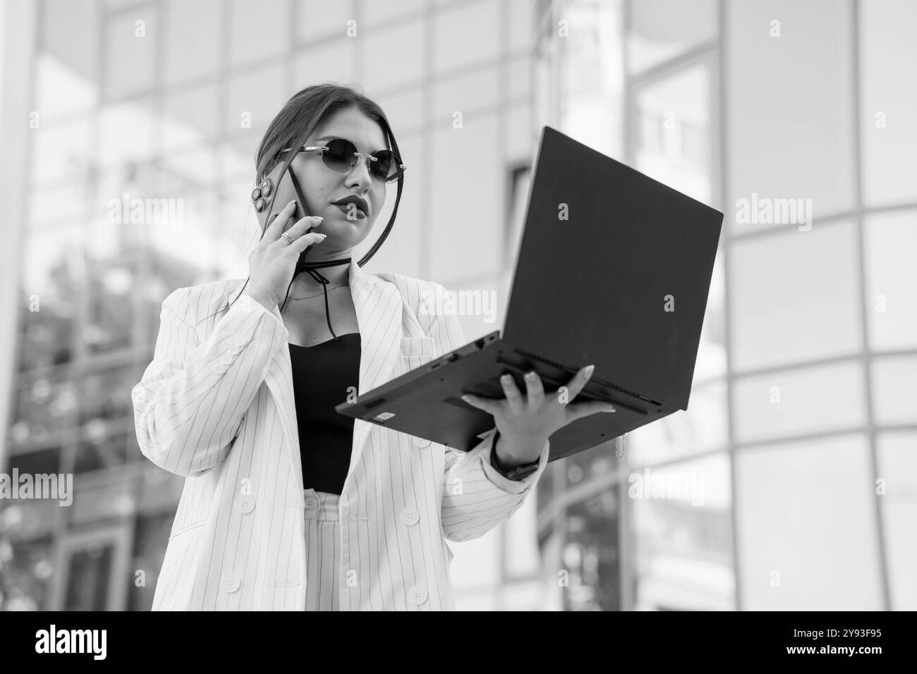 Eine selbstbewusste Geschäftsfrau in einem stilvollen Nadelstreifenanzug mit Laptop im Freien, während sie mit modernem urbanem Hintergrund telefoniert. Stockfoto