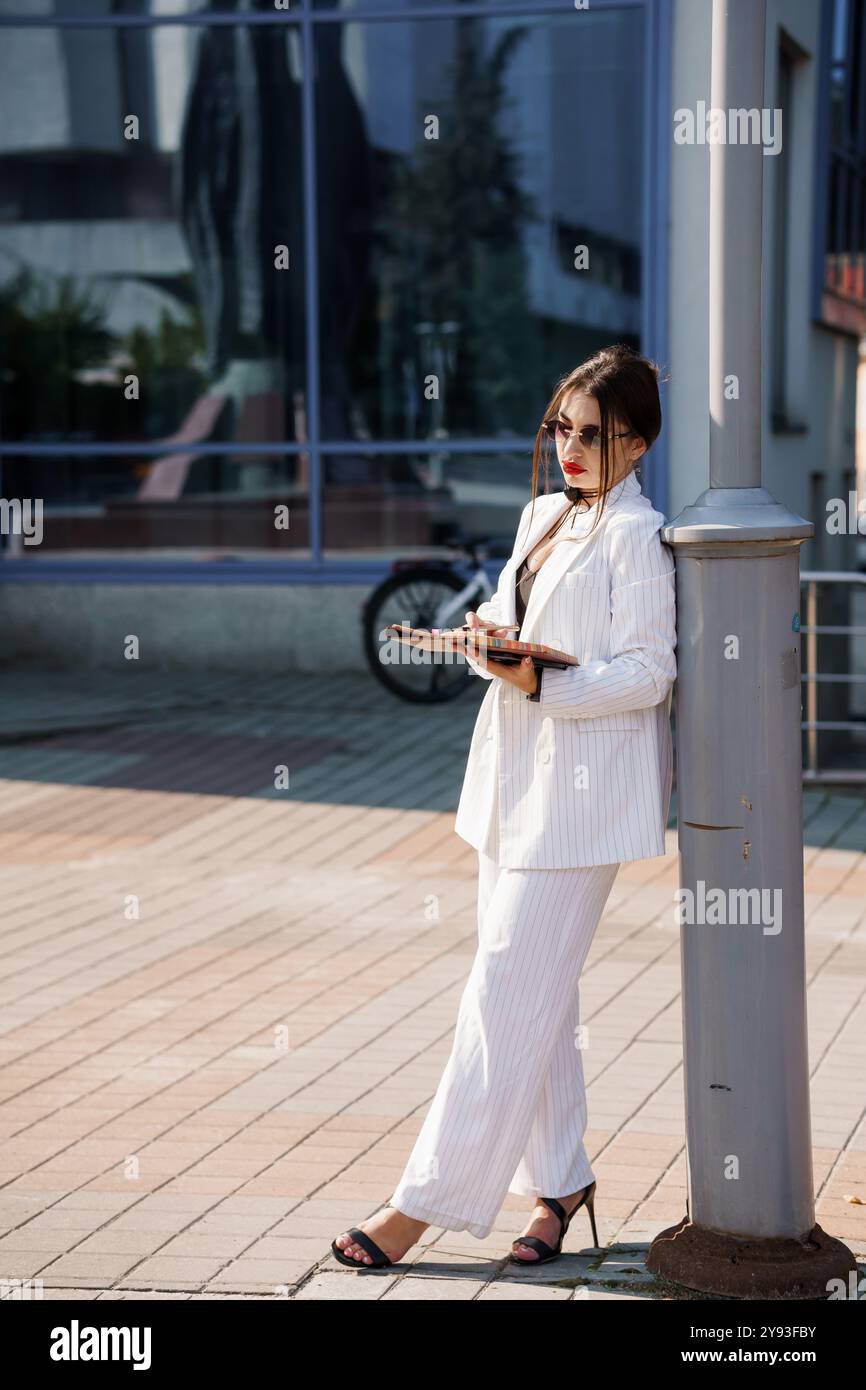 Elegante Geschäftsfrau in stilvollem Nadelstreifenanzug, die selbstbewusst vor einem Pole in urbanem Ambiente posiert. Stockfoto