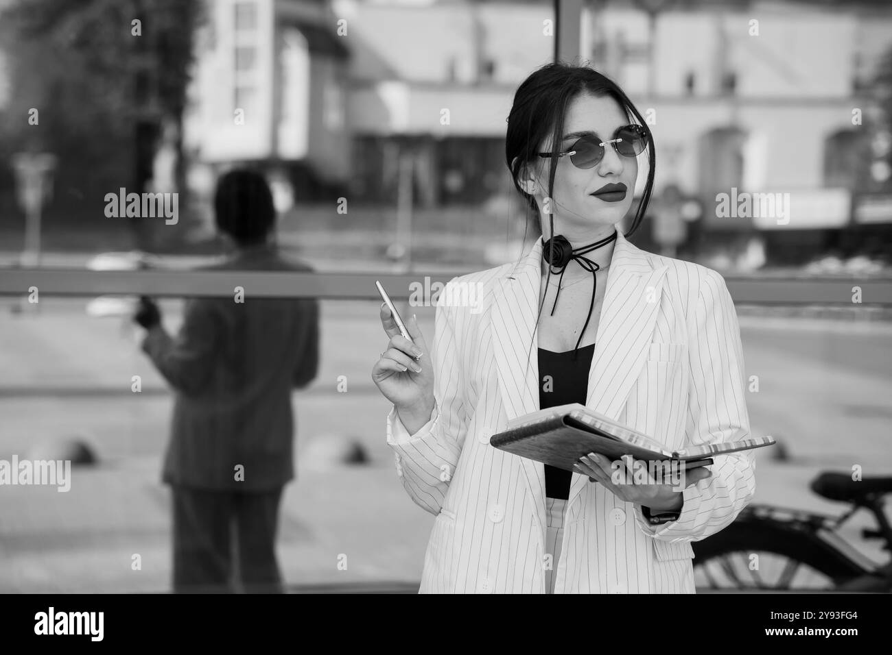 Stilvolle Geschäftsfrau in einer Sonnenbrille, die ein Tablet und einen Stift hält, während sie in der Nähe einer Glasspiegelung steht. Stockfoto