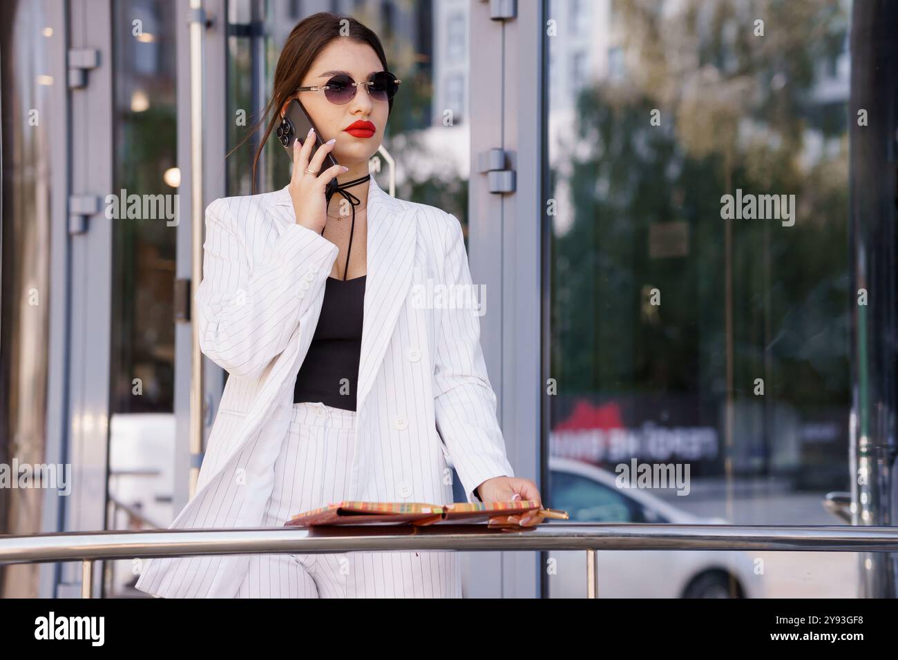 Stilvolle Geschäftsfrau in gestreiftem Anzug, die sich in Telefongesprächen vor dem modernen Bürogebäude engagiert. Stockfoto