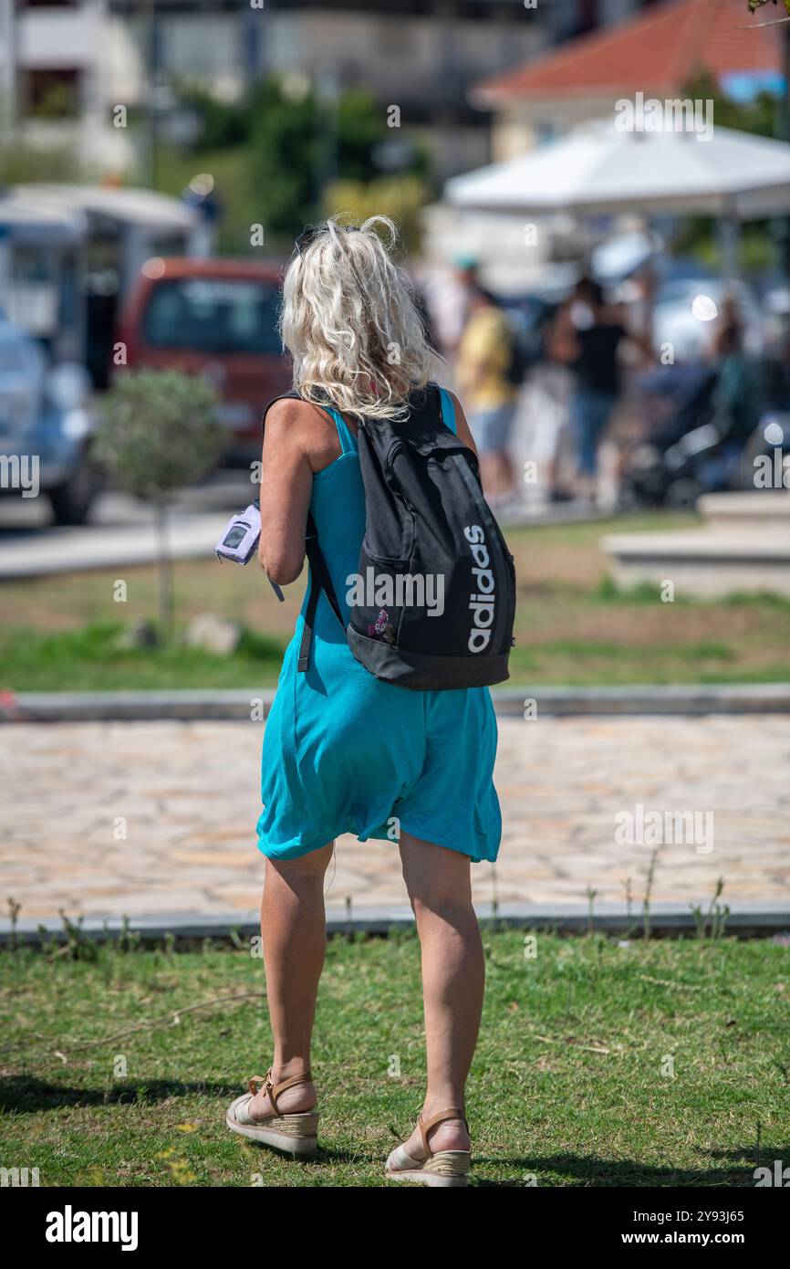 Frau mittleren Alters mit Rucksack und türkisfarbenem Sommerkleid, die allein in einem geschäftigen Stadtzentrum über Gras spaziert. Stockfoto