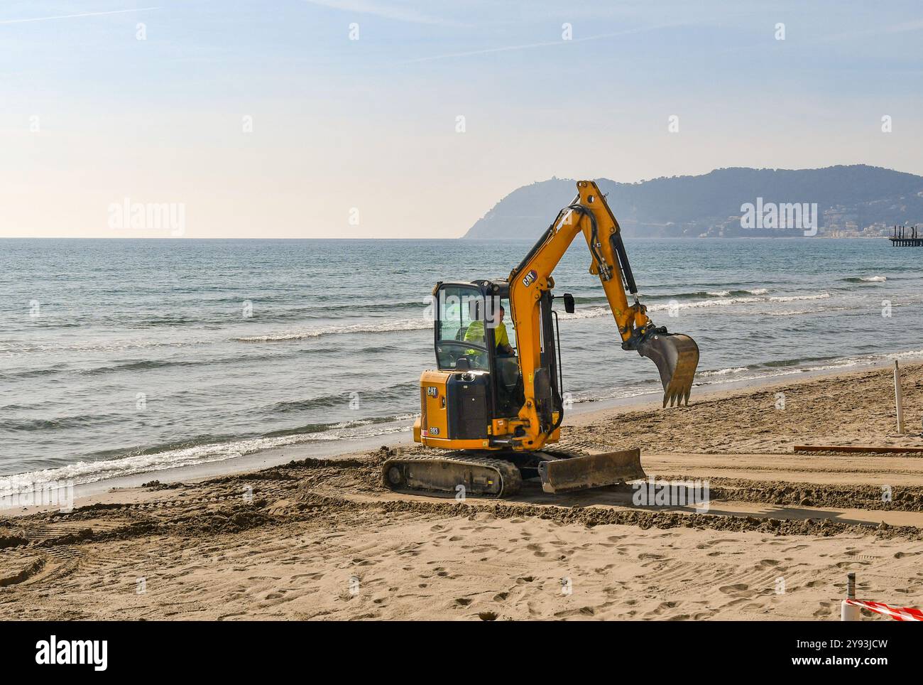 Ein Minibagger der Kategorie 303, der im Frühjahr den Sand des Strandes mit der Landzunge Capo Mele im Hintergrund anordnet, Alassio, Savona, Ligurien, Italien Stockfoto