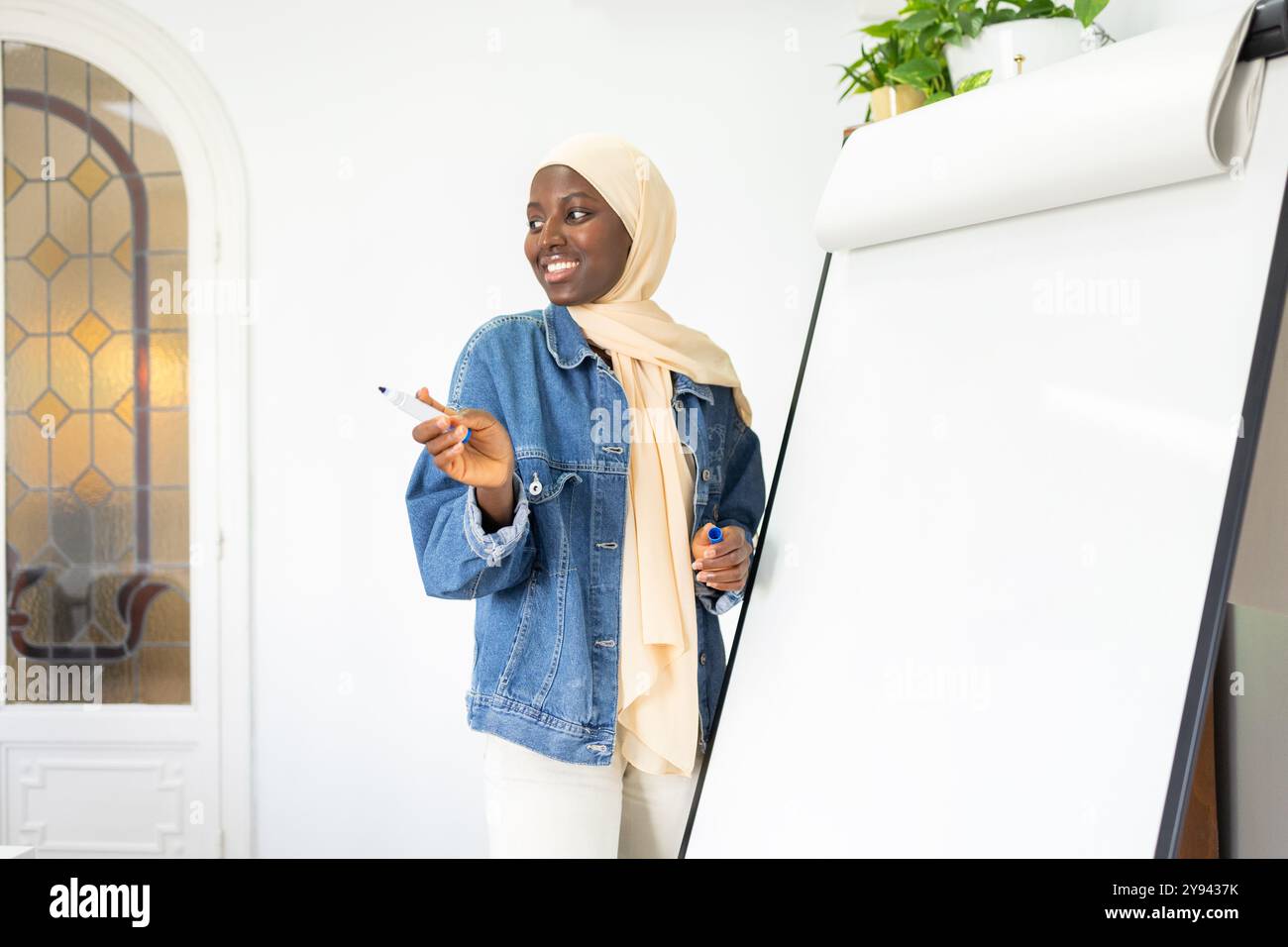 Eine lächelnde schwarze Ärztin in einer Jeansjacke und Hijab-Gesten mit einem Marker neben einem Whiteboard in einem hellen Raum, was auf einen fesselnden Vortrag hindeutet Stockfoto