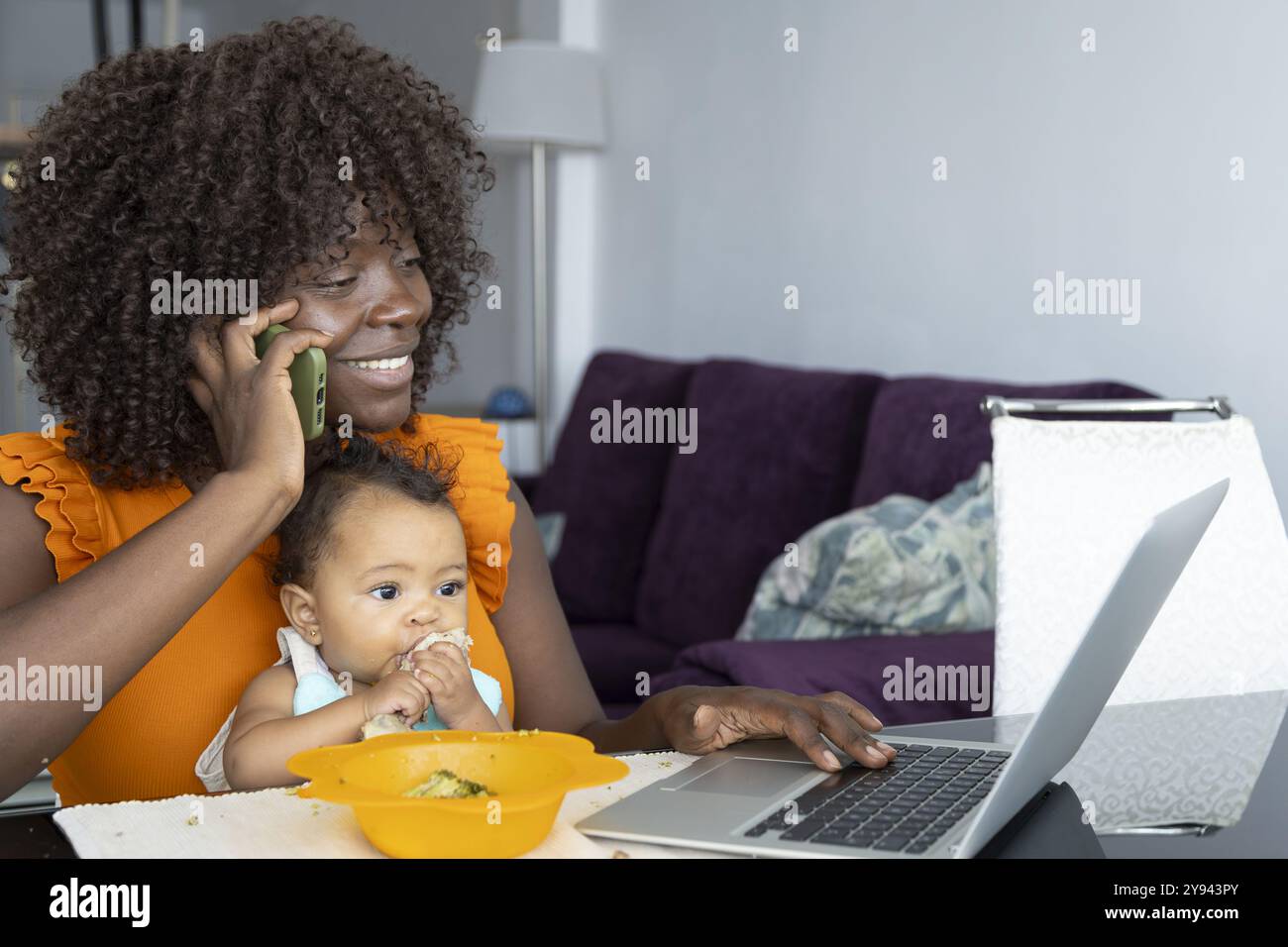 Fröhliche afrikanische Mutter, die einen Laptop benutzt, während sie ihr Baby füttert und zu Hause telefoniert Stockfoto