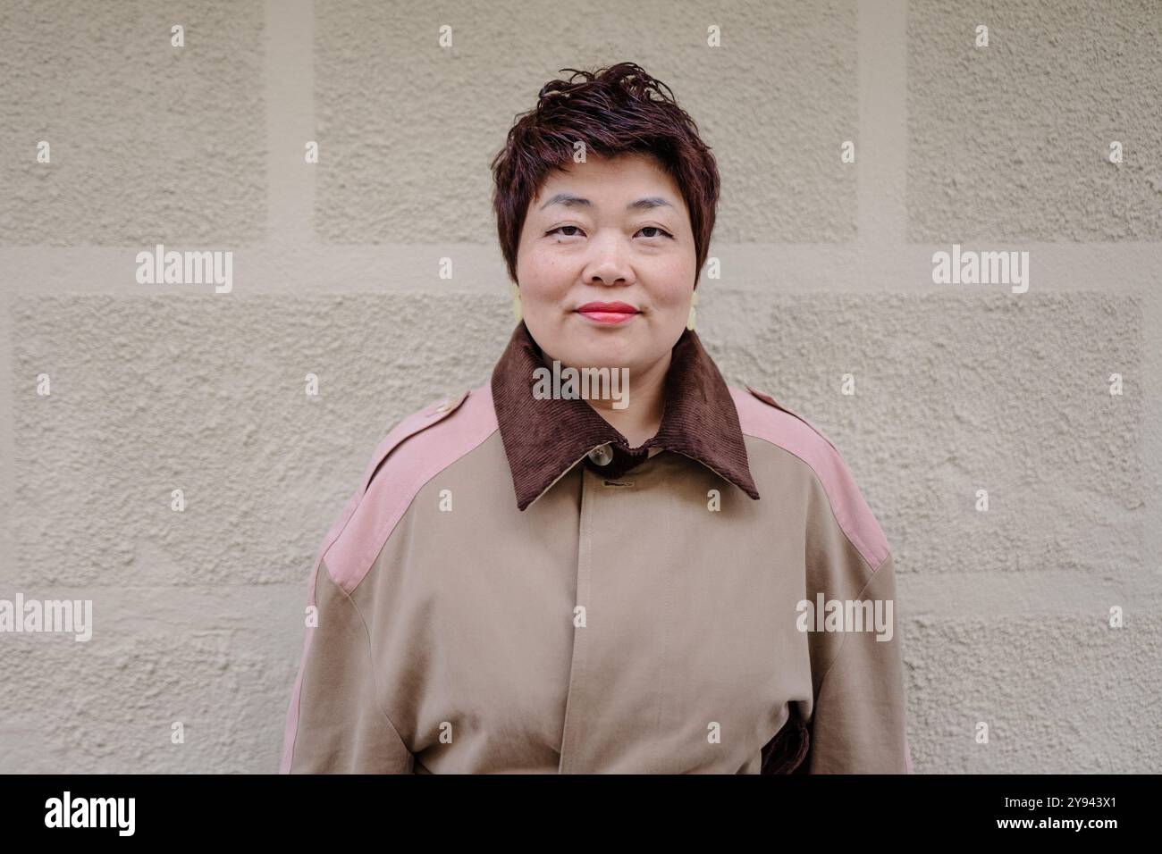 Eine lächelnde, reife asiatische Frau steht selbstbewusst vor einer strukturierten beigefarbenen Wand. Ihr stylischer, mehrfarbiger Mantel mit braunem Kragen trägt zu ihrem Cheerf bei Stockfoto