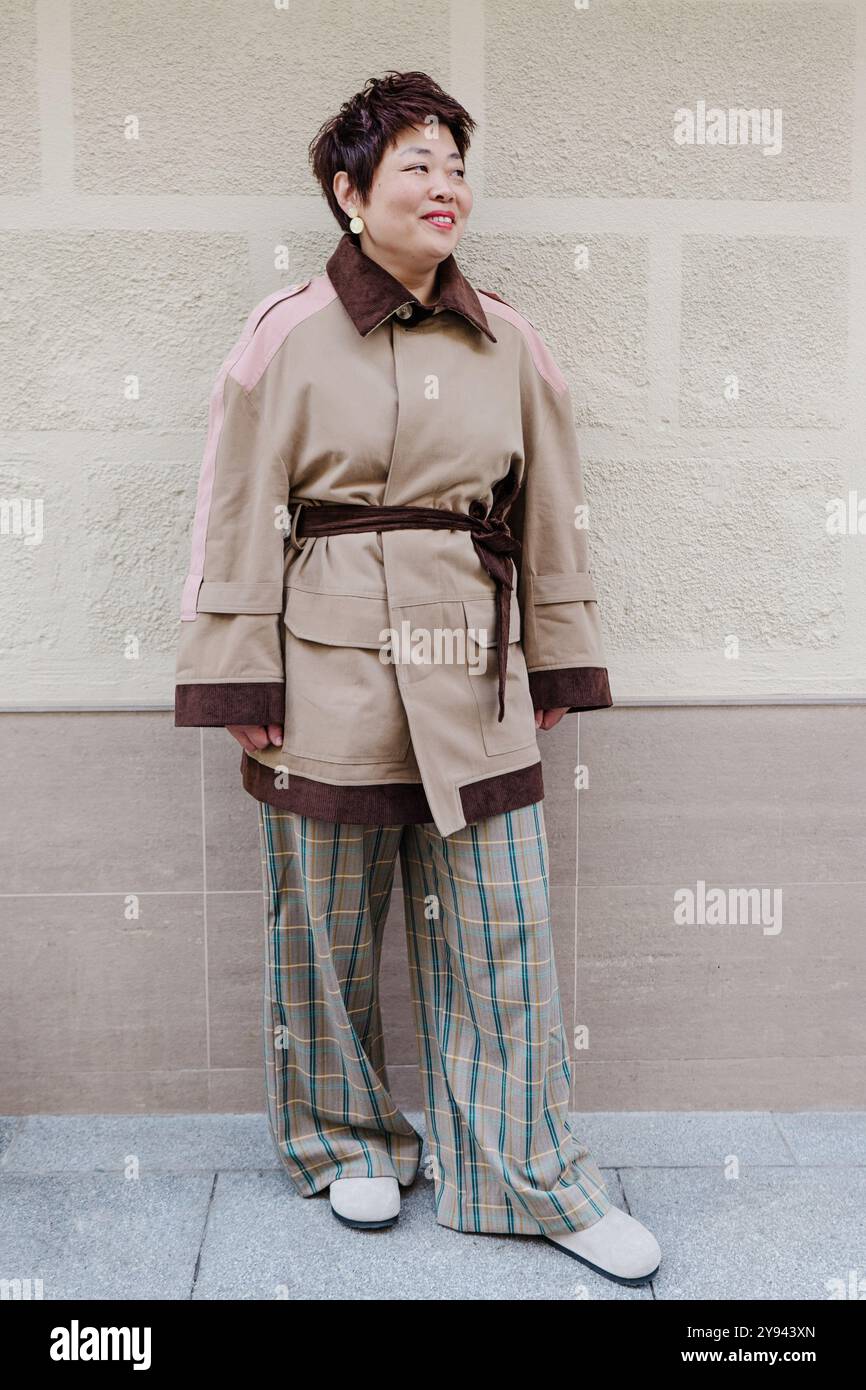 Asiatische Frau mittleren Alters in einem modischen Trenchcoat mit rosa Akzenten und karierten Hosen, die selbstbewusst vor einem hellen Hintergrund lächelt Stockfoto