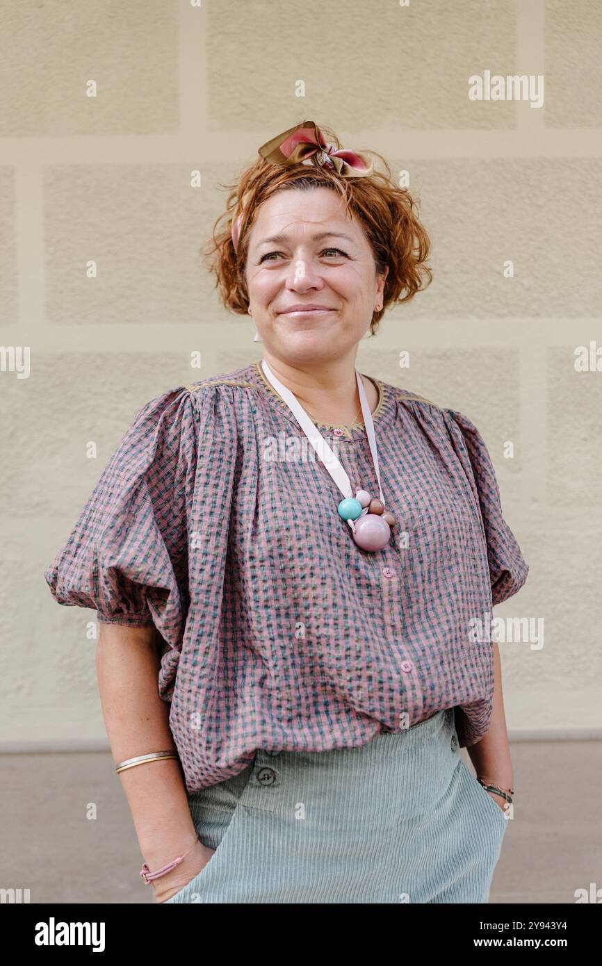 Ein Porträt einer lächelnden Frau mittleren Alters in einem stilvollen Outfit, die selbstbewusst vor einer hellen Ziegelwand steht Stockfoto