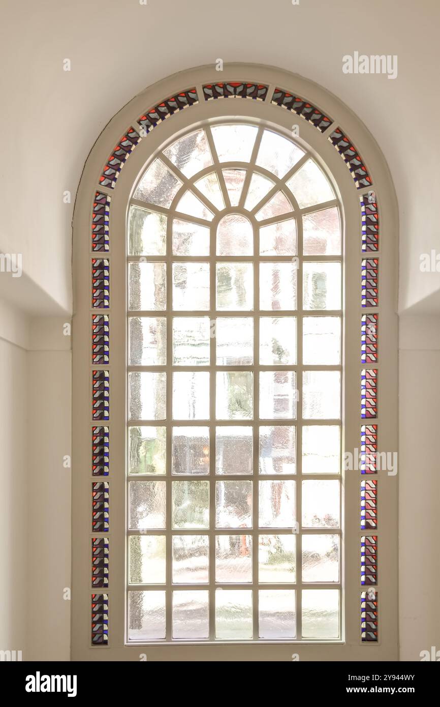 Ein hohes Bogenfenster mit Buntglasdetails an den Kanten, das Sonnenlicht in einen hellen Raum mit neutralen Wänden lässt. Stockfoto