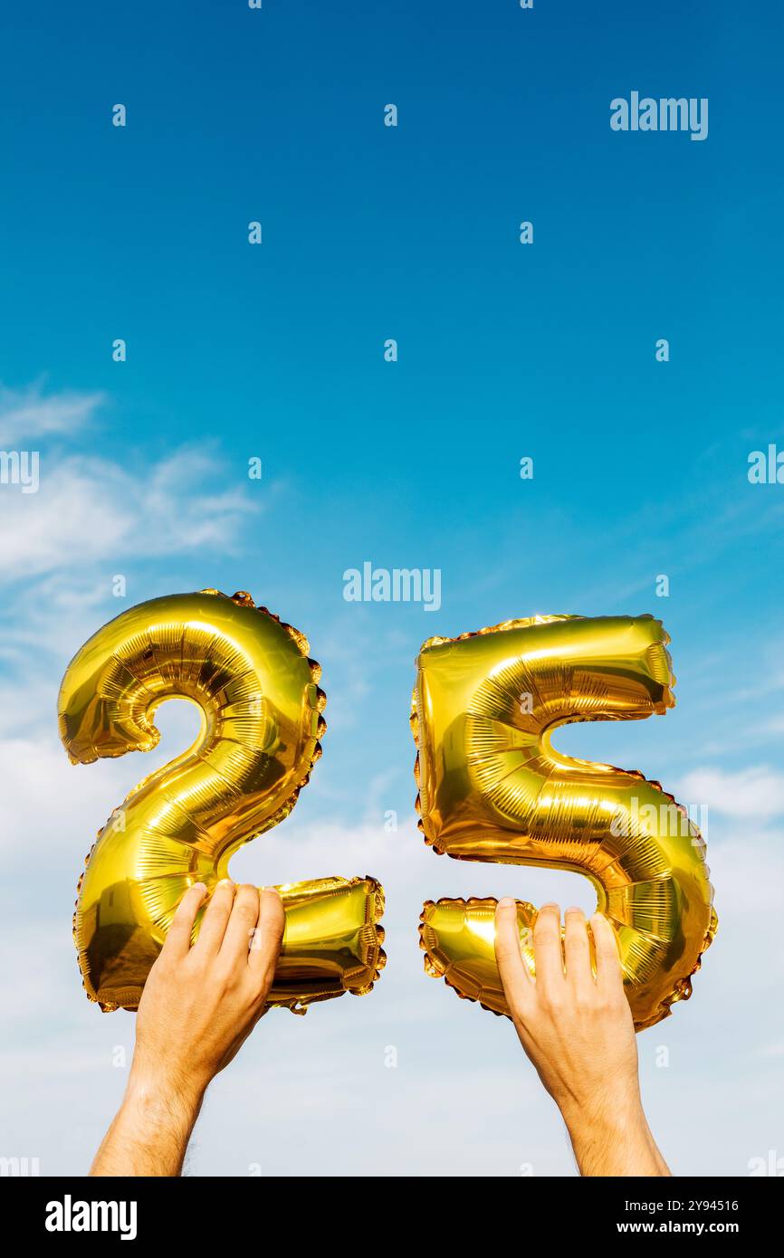Beschnittene Ansicht der Hände, die glänzende goldene Ballons mit der Nummer 25 halten, mit einem klaren blauen Himmel als Hintergrund. Stockfoto
