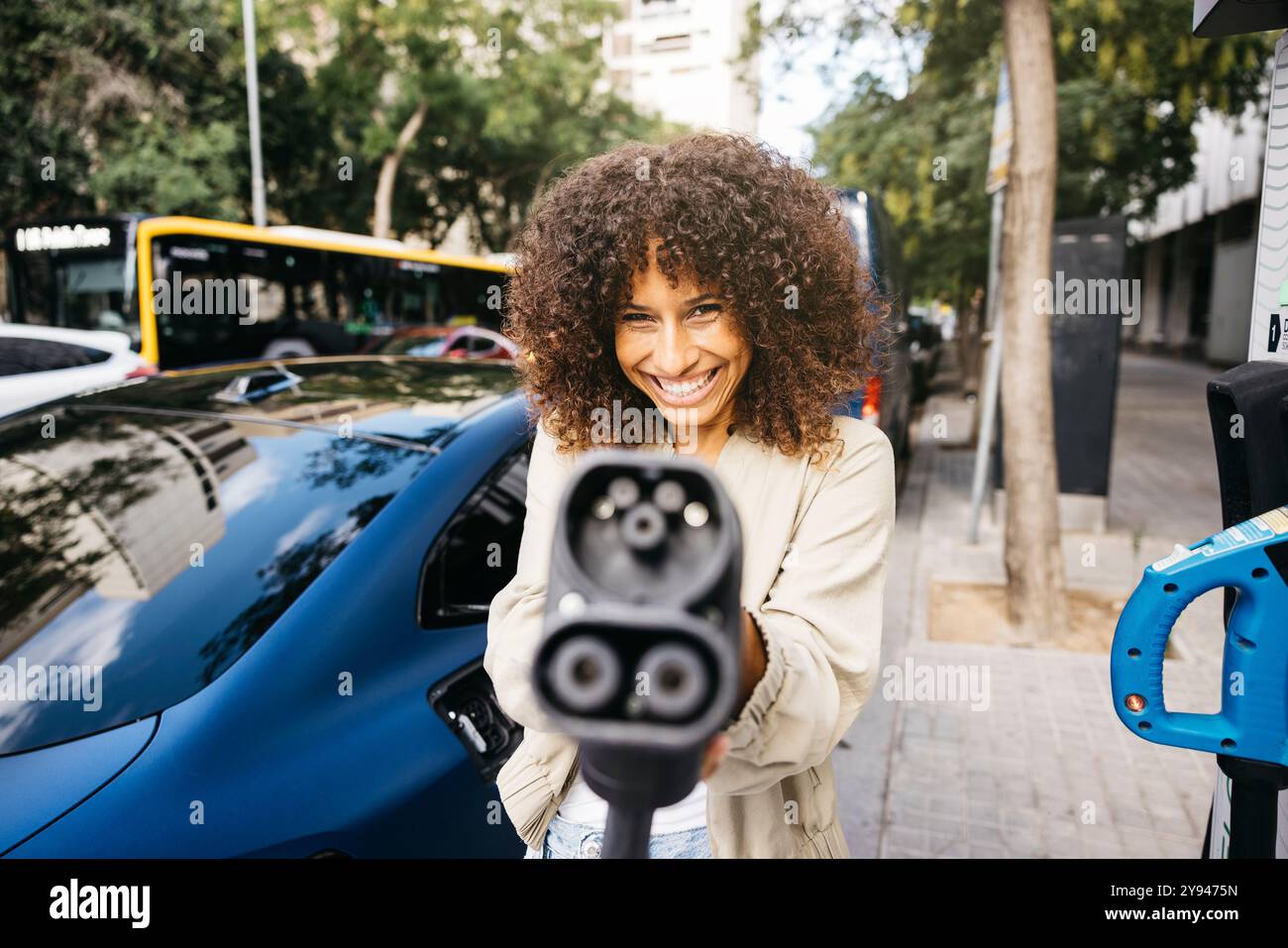 Eine freudige Frau, die mit dem EV-Ladegerät spielt, es als Waffe benutzt, während sie ihr Elektroauto an einer Ladestation in der Stadt auflädt. Stockfoto