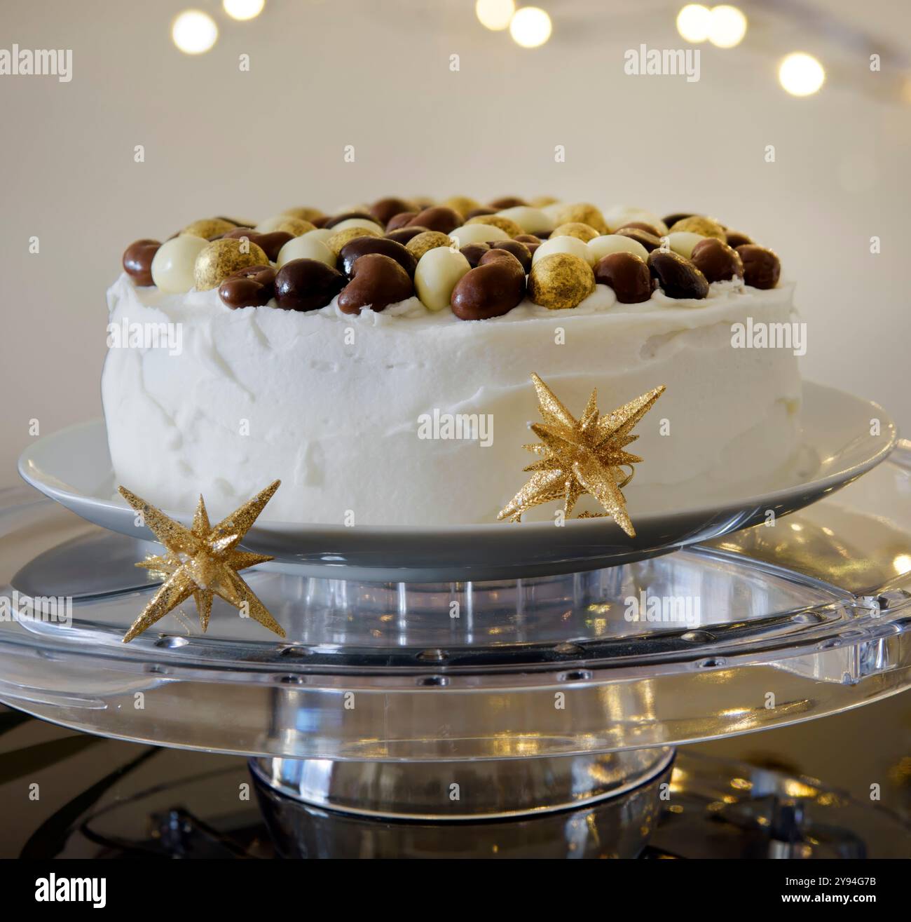 Eisgekühlter Weihnachtskuchen mit Nüssen mit Schokoladenüberzug auf einem klaren Ständer mit unscharfen Weihnachtslichtern im Hintergrund Stockfoto