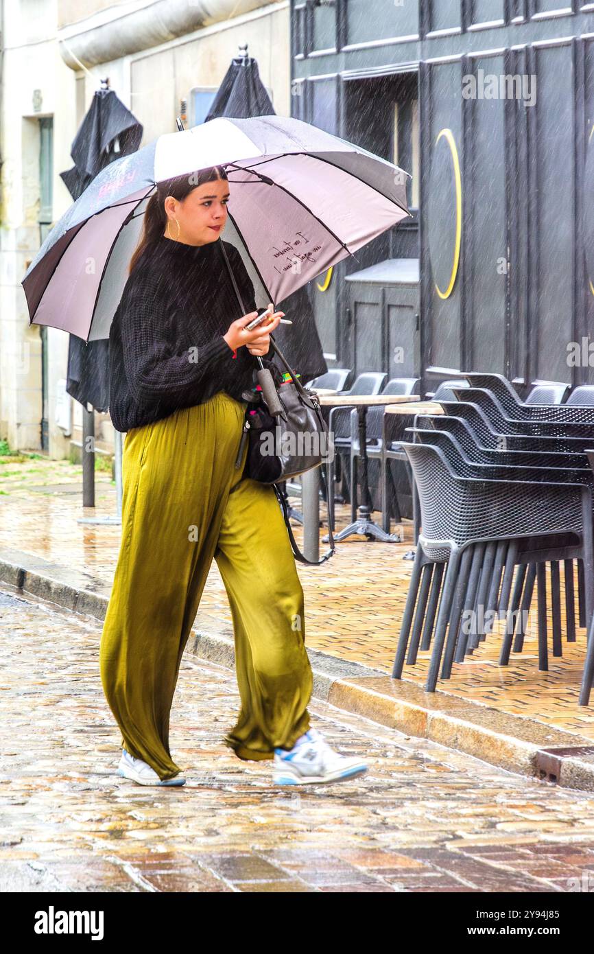 Frau in Baggy Hosen mit Regenschirm, die im strömenden Regen die Straße im Stadtzentrum entlang läuft - Tours, Indre-et-Loire (37), Frankreich. Stockfoto