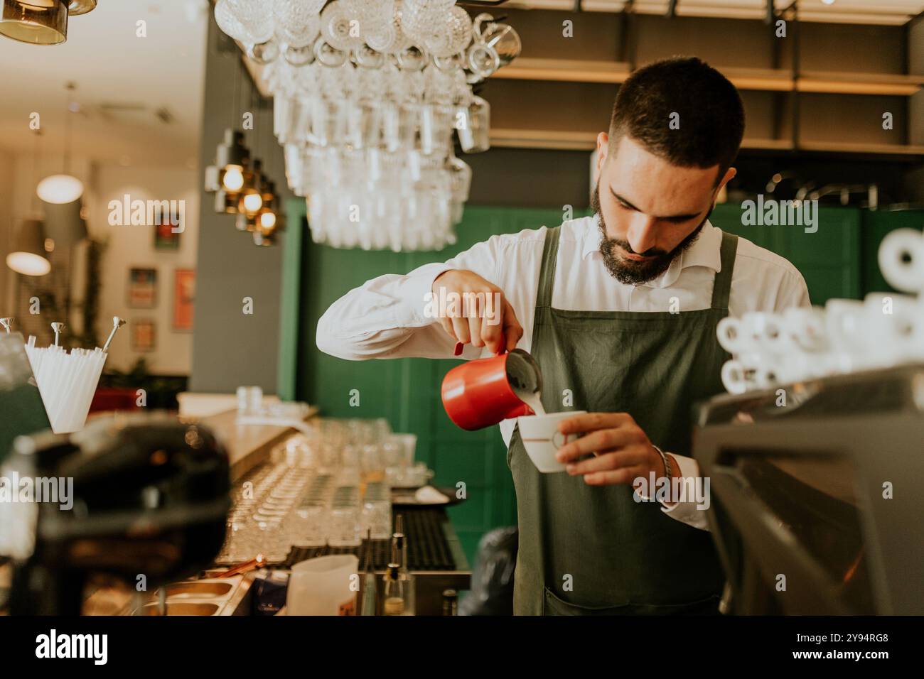 In einem warmen und einladenden Café gießt ein engagierter Barista fachmännisch Milch in eine Kaffeetasse und sorgt so für ein angenehmes Erlebnis für Gäste. Stockfoto