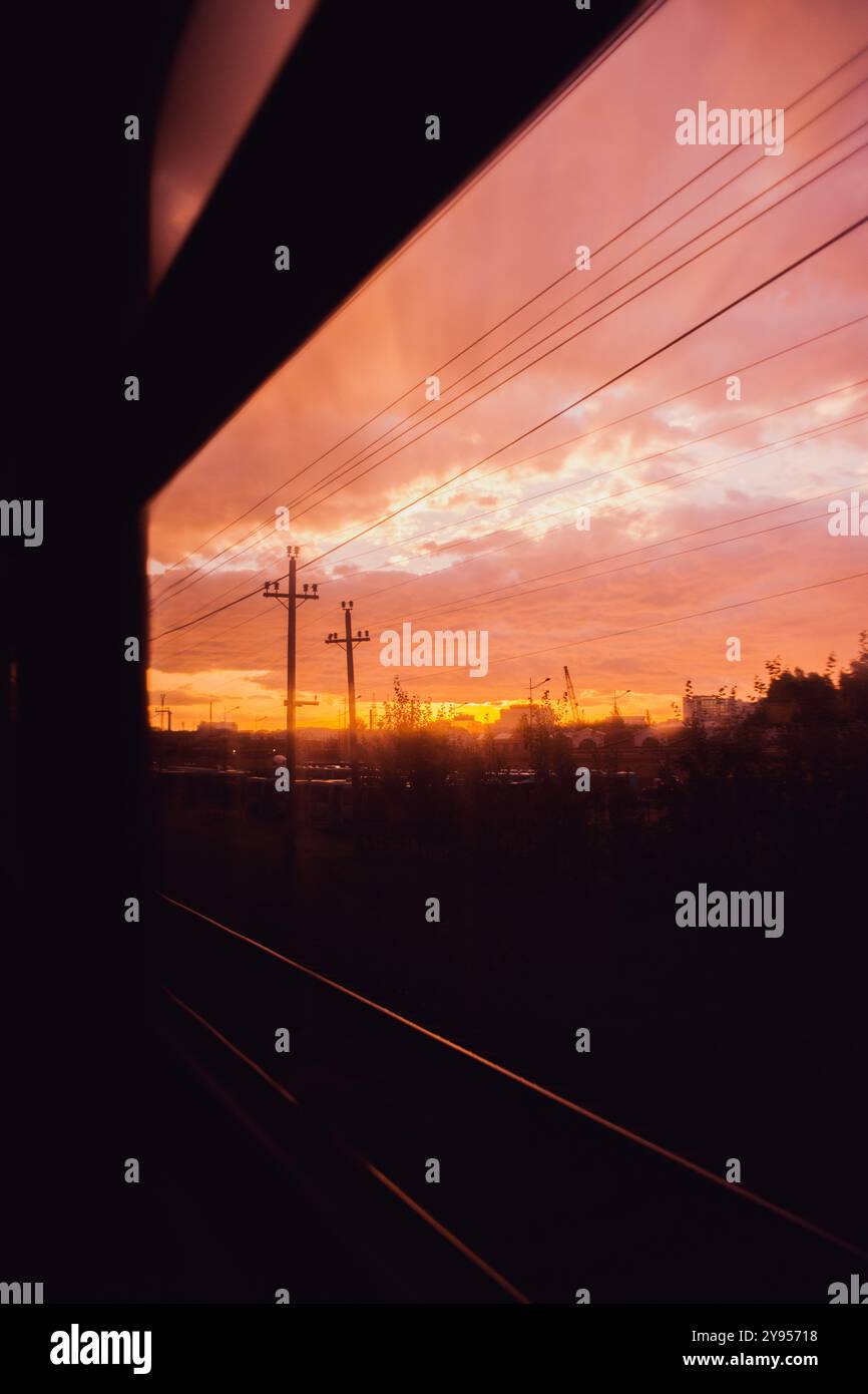 Durch das Fenster eines fahrenden Zuges ist ein heller Sonnenaufgang zu sehen. Es beleuchtet die Stromleitungen, die Schienen und die Silhouette der Stadt mit ihrem Strahl Stockfoto