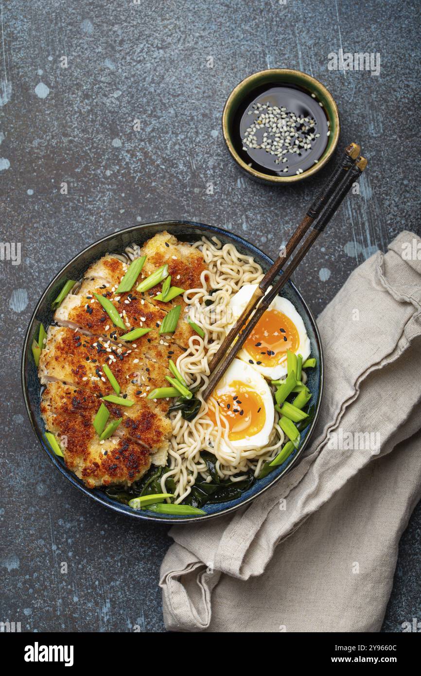 Asiatische Nudeln Ramen-Suppe mit frittiertem Panko-Hähnchenfilet und gekochten Eiern in einer Keramikschüssel mit Koteletts und Sojasauce auf rustikalem Backgroun aus Stein Stockfoto