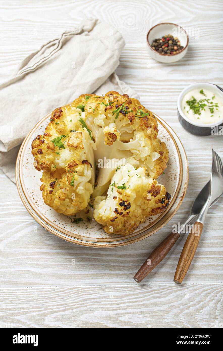 Ganzer gerösteter Blumenkopf mit Olivenöl auf Teller auf weißem hölzernem, rustikalem Tisch mit Besteck. Vegetarisches Gericht, Lebensmittelfotografie, Essen Stockfoto